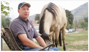 man sitting in chair talking to camera with horse behind him